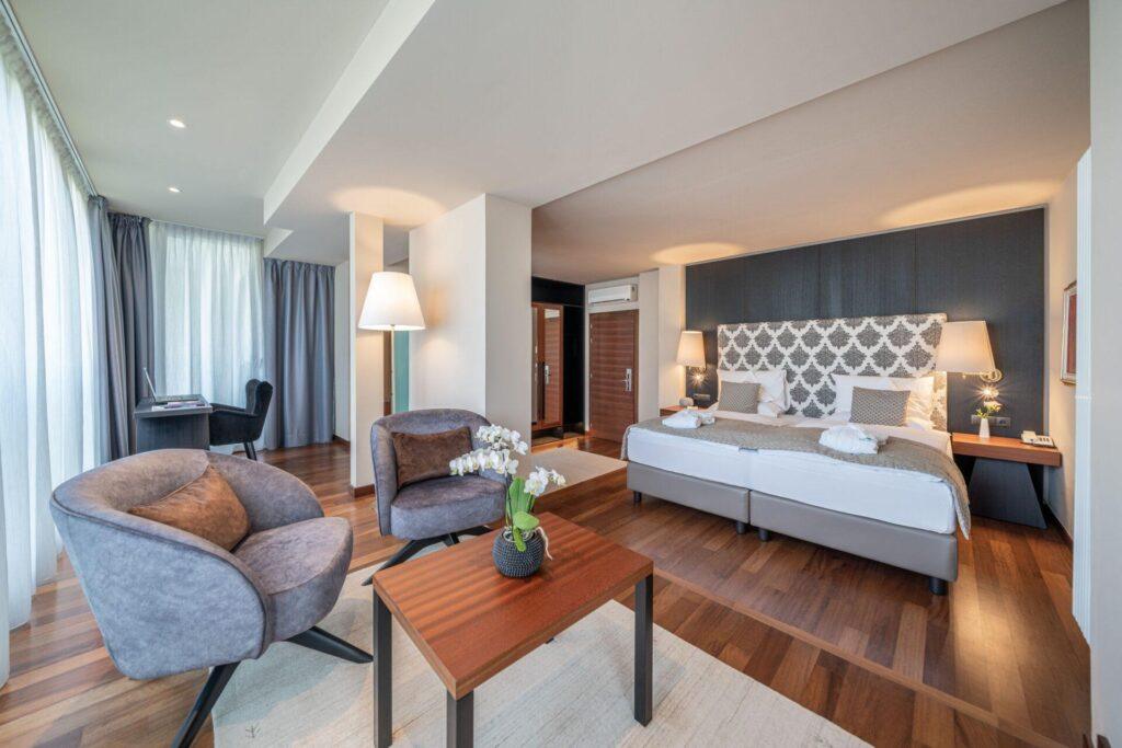Geräumiges Hotelzimmer mit einem großen Bett, einer Sitzecke mit zwei Stühlen und einem Tisch, einem Schreibtisch am Fenster, Holzböden und moderner Einrichtung.