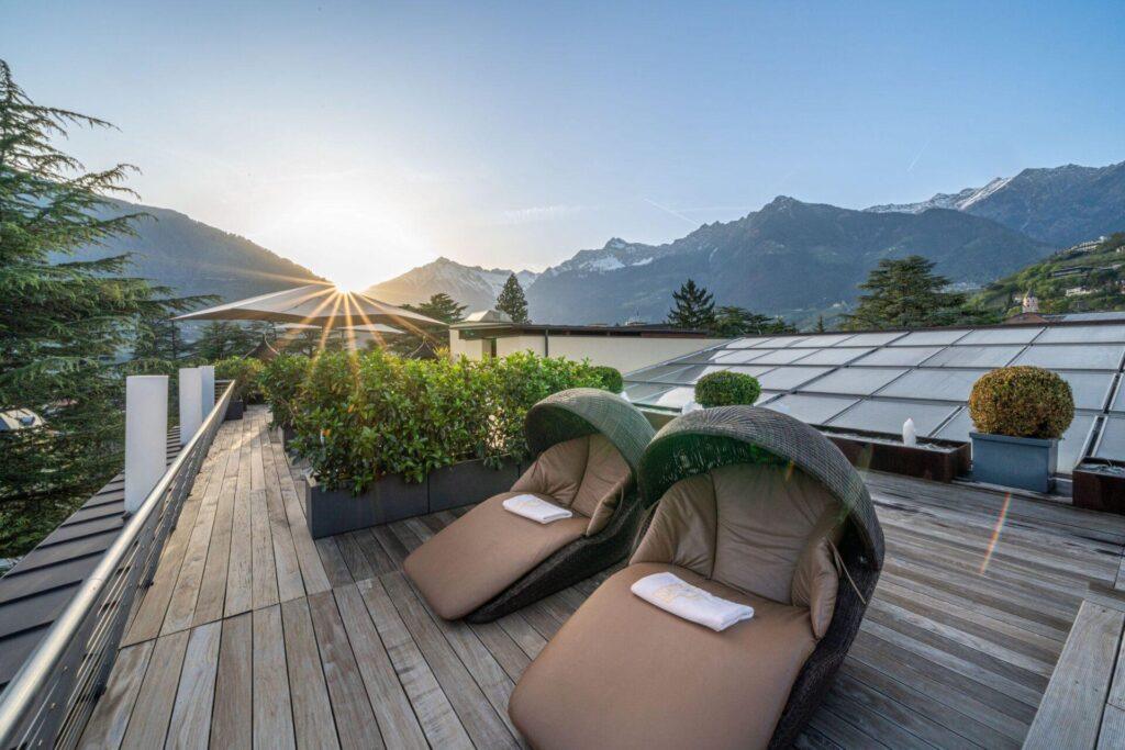 Zwei Liegestühle auf einer Holzterrasse mit Blick auf die Berge und den Sonnenaufgang, umgeben von Topfpflanzen und Grünpflanzen.