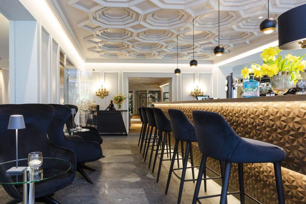 Moderne Hotellounge mit dunkelblauen Sesseln, hohen Barhockern, geometrischer Barfront, dekorativer Decke und Vasen mit gelben Blumen auf dem Tresen.