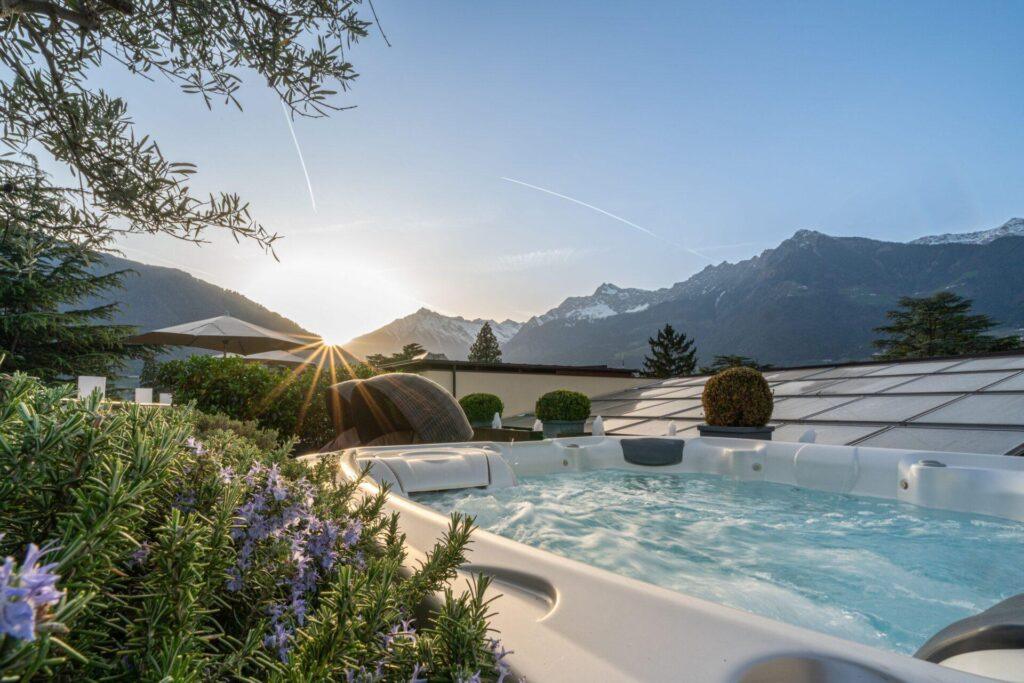 Whirlpool im Freien, umgeben von Grün, mit den Bergen im Hintergrund und der Sonne, die hinter den Gipfeln untergeht.