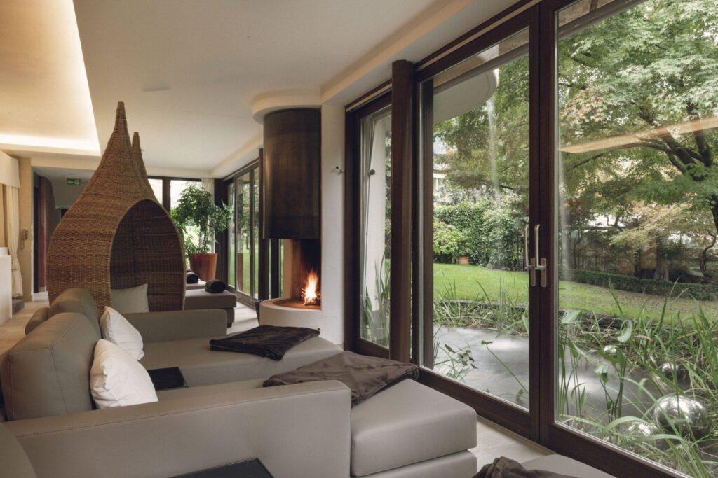 Ein moderner Ruhebereich mit beigen Sofas, einem Korbsessel, einem kleinen Kamin und großen Glastüren mit Blick auf einen grünen Garten.