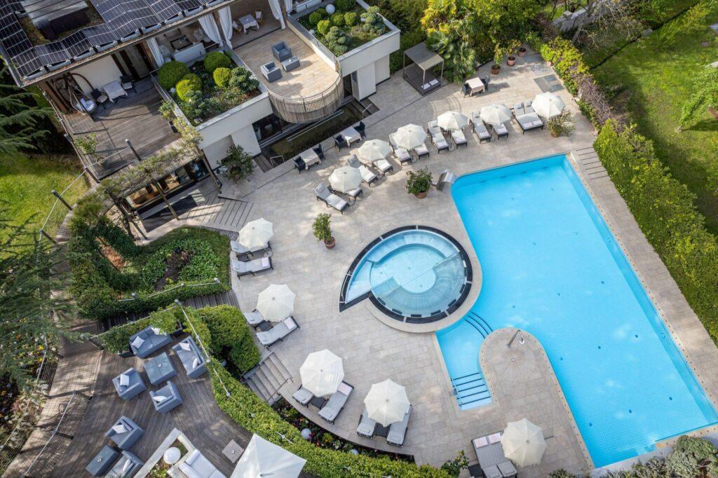 Luftaufnahme eines modernen Außenpools mit Liegestühlen, Sonnenschirmen, einem runden Whirlpool, Sitzplätzen auf der Terrasse und einer Grünanlage, die ein großes Gebäude umgibt.