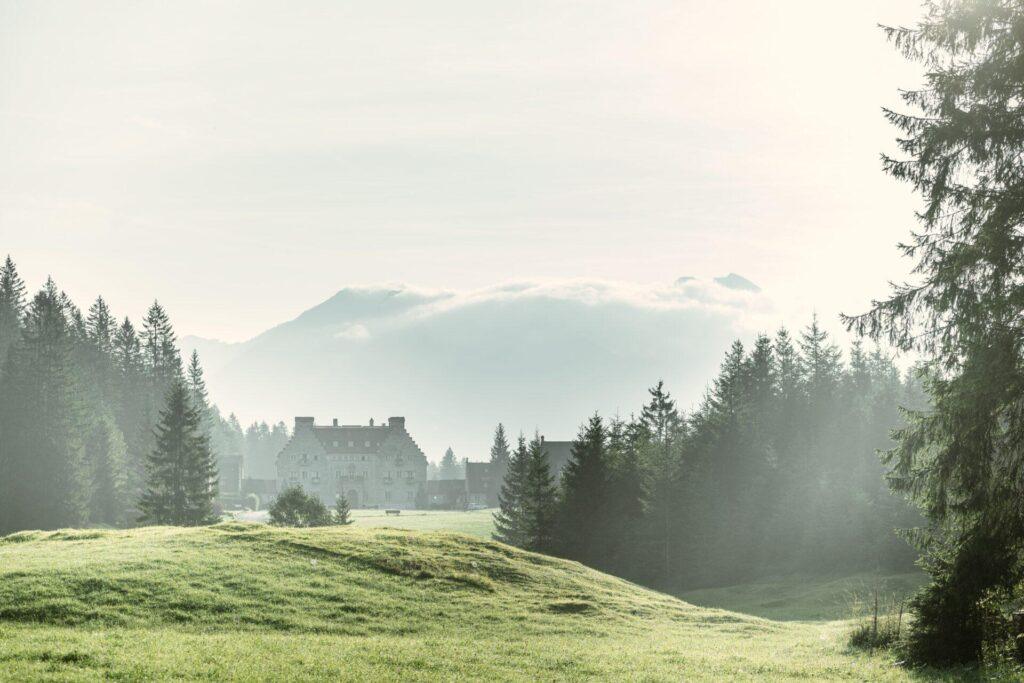 Das große Haus DAS KRANZBACH liegt auf einer grasbewachsenen Lichtung, umgeben von Kiefern, mit den nebelverhangenen bayerischen Alpen und einem wolkenverhangenen Himmel im Hintergrund - ein einladender Rückzugsort inmitten einer atemberaubenden Natur.