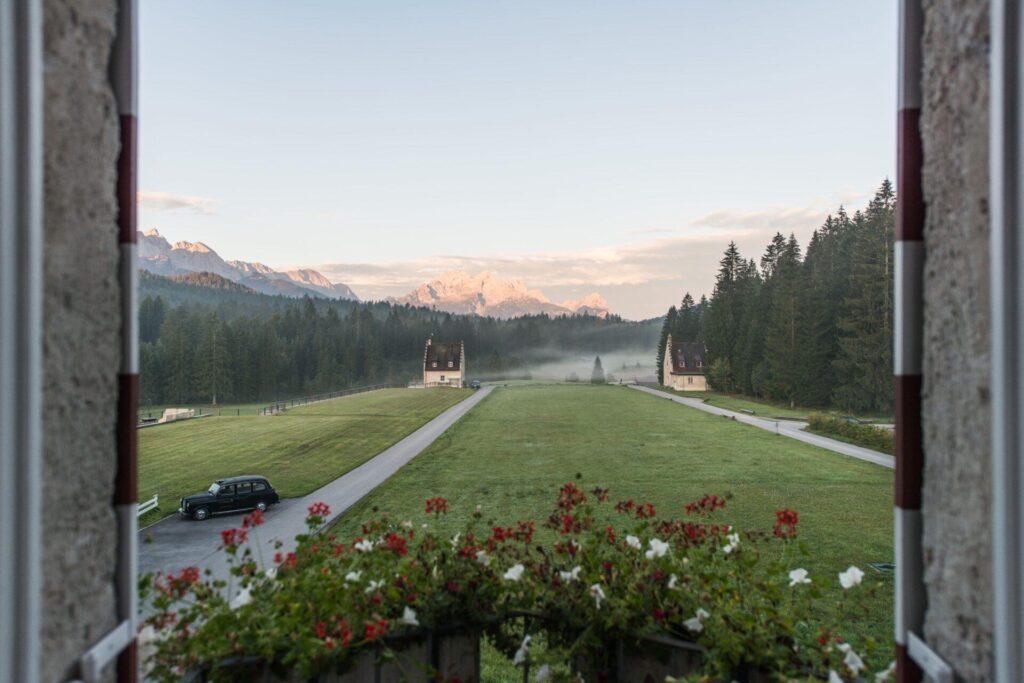 Blick aus dem Fenster auf eine Wiese mit zwei Häusern, einem geparkten Auto, Bäumen auf beiden Seiten und den majestätischen bayerischen Alpen im Hintergrund - nur wenige Augenblicke entfernt von der Ruhe des KRANZBACH Retreat.