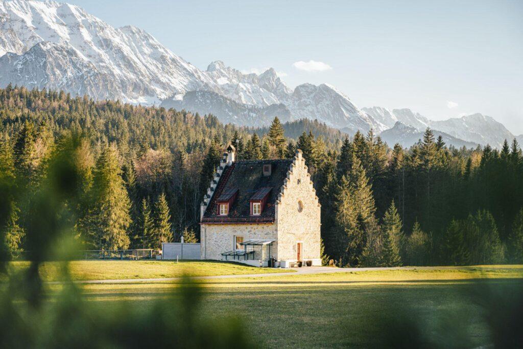 Ein steinernes Haus mit steilem Dach steht auf einer Wiese, umgeben von Wald, mit den schneebedeckten Gipfeln der bayerischen Alpen im Hintergrund - ein idyllisches Refugium, das an DAS KRANZBACH erinnert.