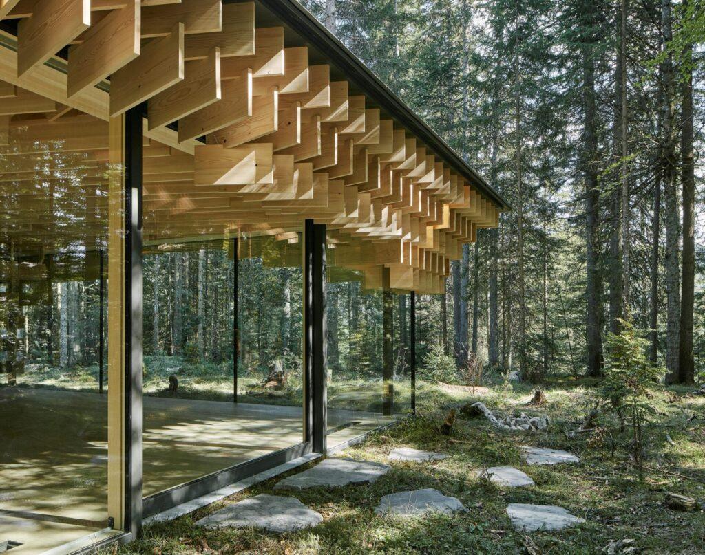 Modernes Glasgebäude mit geometrischem Holzüberbau, eingebettet in einen Wald in der Nähe der bayerischen Alpen, mit Pflastersteinen, die zum Eingang des KRANZBACH Retreats führen.