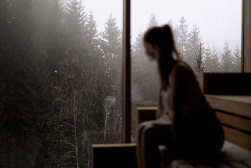 Ein Mensch sitzt auf einer Holzbank im Innenbereich von DAS KRANZBACH und blickt aus einem großen Fenster auf einen nebligen Wald und die majestätischen bayerischen Alpen - ein perfekter Rückzugsort für ruhige Besinnung.