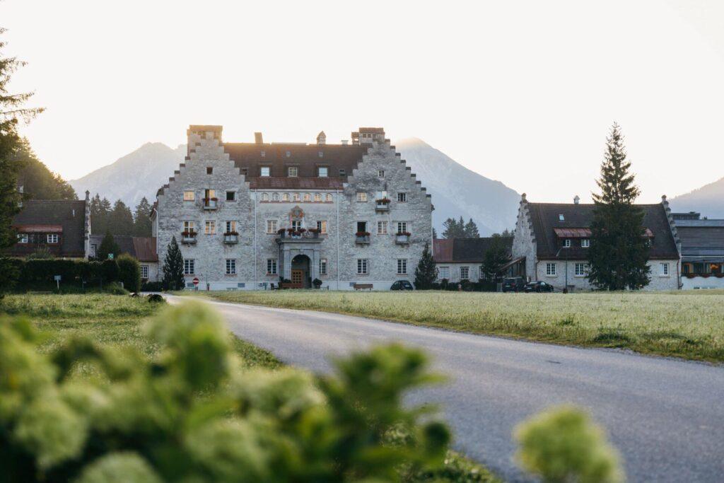 Ein großes Steingebäude mit Stufengiebel steht am Ende einer gepflasterten Straße und bietet einen ruhigen Rückzugsort in den bayerischen Alpen, mit Bergen und Bäumen im Hintergrund.
