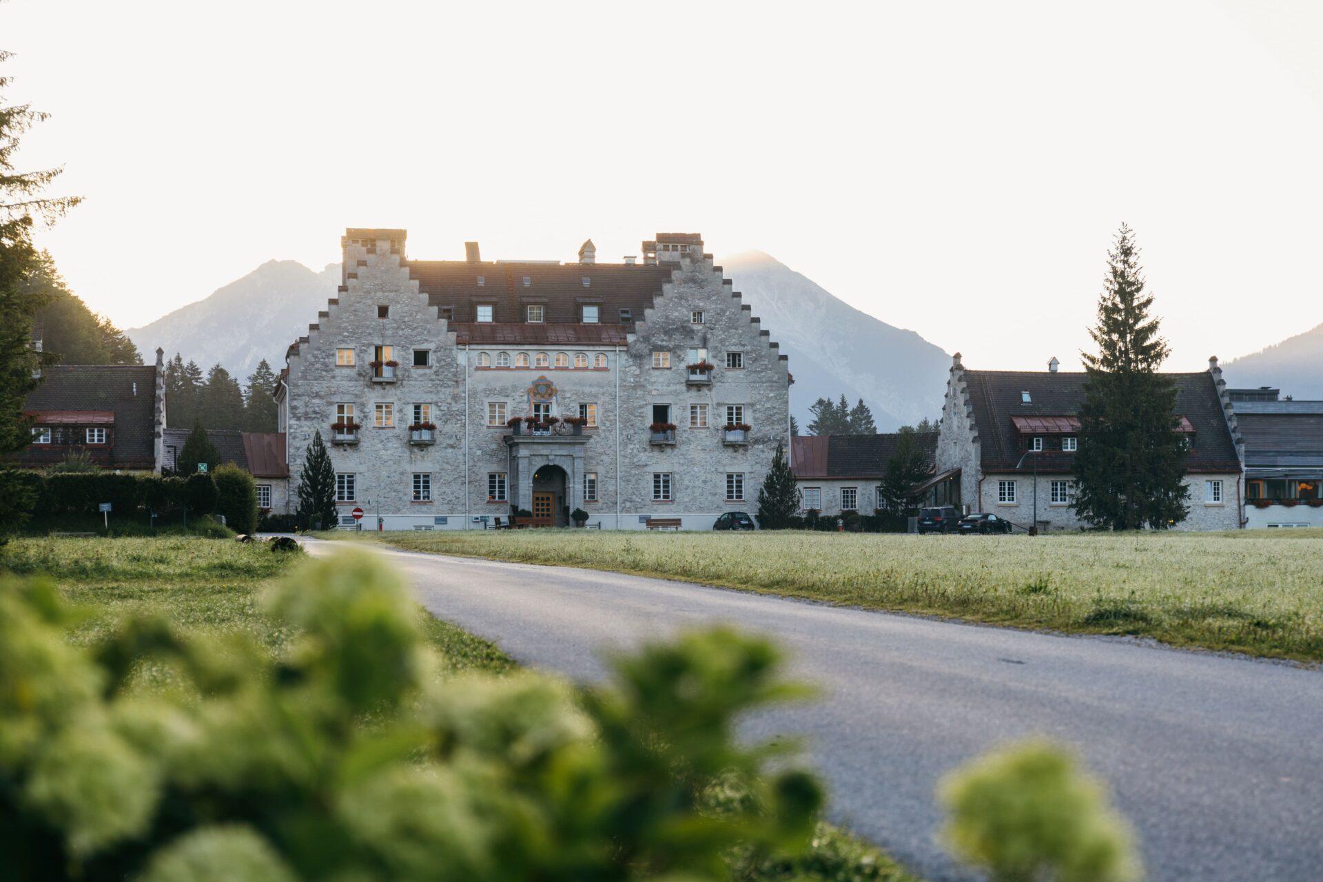 Ein großes Steingebäude mit Stufengiebel steht am Ende einer gepflasterten Straße und bietet einen ruhigen Rückzugsort in den bayerischen Alpen, mit Bergen und Bäumen im Hintergrund.