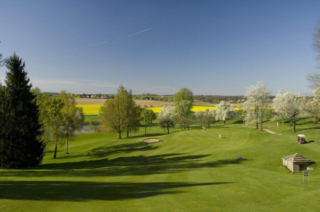 Ein gut gepflegter Golfplatz mit Bäumen, einem Sandbunker und einem kleinen Teich, umgeben von Feldern und einem klaren blauen Himmel.