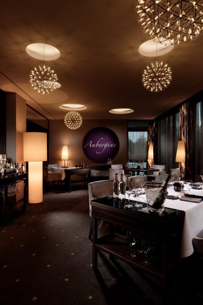 Gehobenes Restaurant mit moderner Beleuchtung, gedeckten Tischen, bequemen Sitzgelegenheiten und einem lila Schild mit der Aufschrift "Aubergine" an der Rückwand.