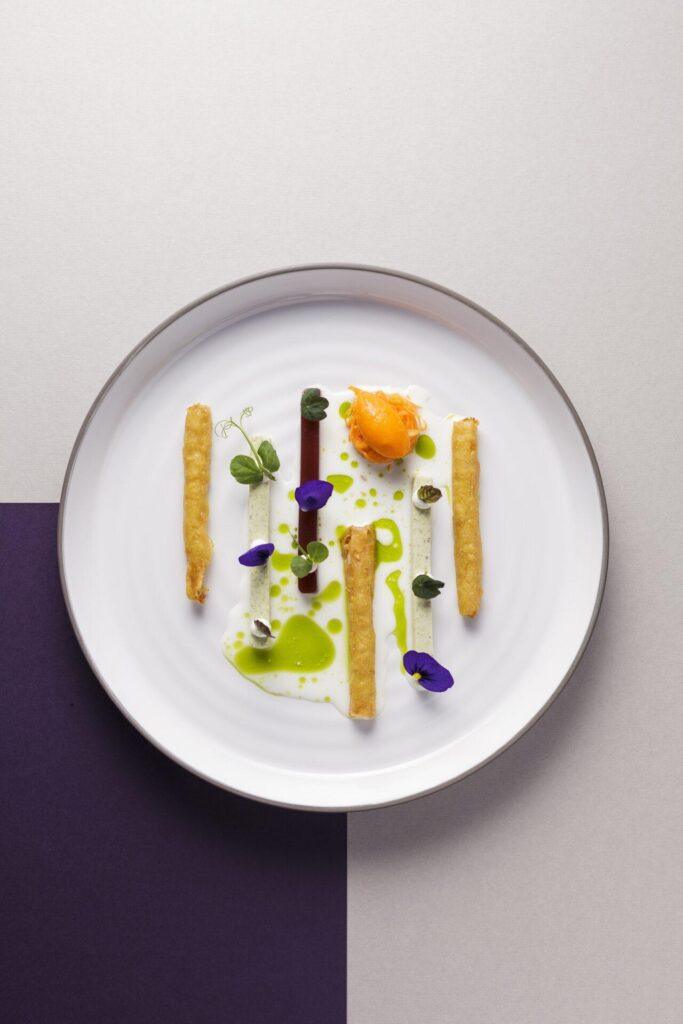 Ein weißer Teller mit vier panierten Stäbchen, grüner Soße, weißer Sahne, essbaren Blumen, Kräutern und einem orangefarbenen Klecks, angeordnet in einem minimalistischen Stil auf einem zweifarbigen Hintergrund.