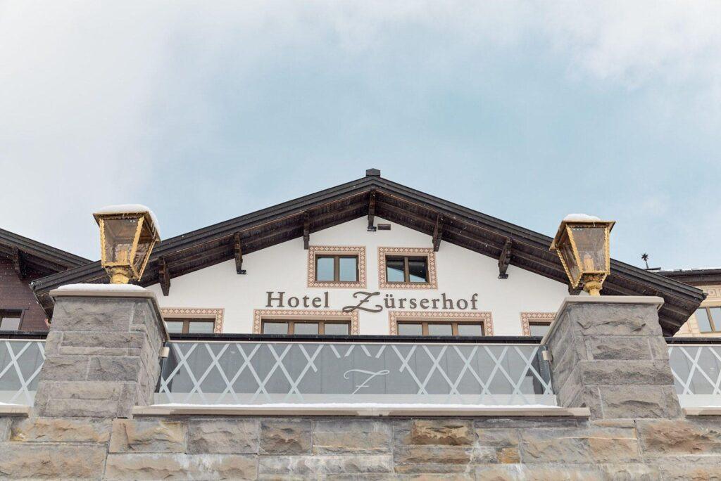 Das Bild zeigt die Frontfassade des Hotels Zürserhof mit Giebeldach, Steinsäulen und goldverzierten Laternen unter einem bewölkten Himmel.