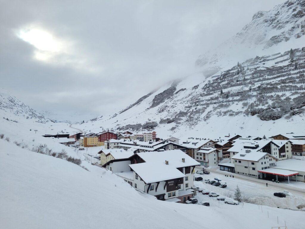 Ein kleines Bergdorf mit schneebedeckten Häusern und Straßen liegt am Fuße eines verschneiten, wolkenverhangenen Hügels unter einem wolkenverhangenen Himmel.