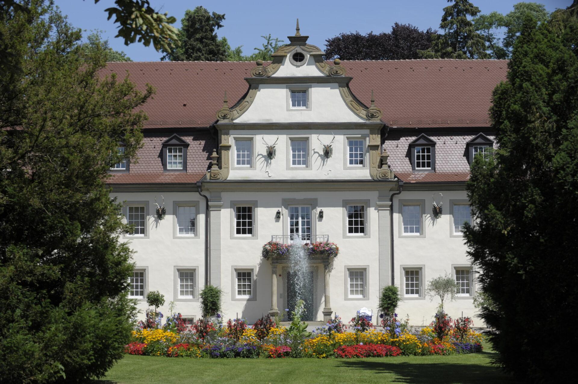 Ein großes, symmetrisches weißes Gebäude mit rotem Ziegeldach, verziert mit Geweihschildern über dem Eingang, umgeben von einem gepflegten Rasen und bunten Blumenbeeten.