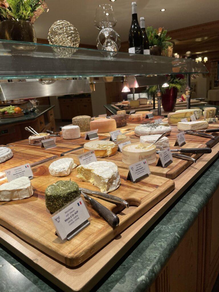 Ein Käsebuffet mit verschiedenen ganzen und in Scheiben geschnittenen Käsesorten auf Holzbrettern, die jeweils beschriftet sind, mit Serviermessern und zwei Weinflaschen im Hintergrund.