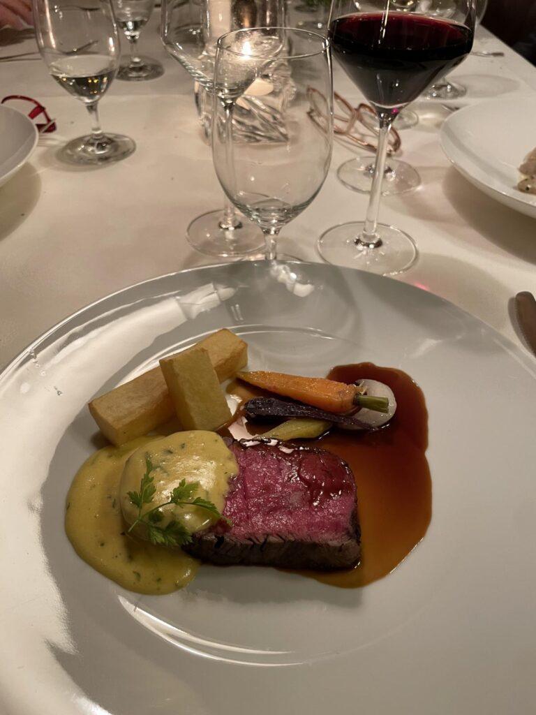 Ein Teller mit einem medium-rare Steak, zwei rechteckigen Kartoffelstücken, Karotten, einem grünen Gemüse, gelber Soße, brauner Soße und einem Glas Rotwein auf einem weißen Tischtuch.