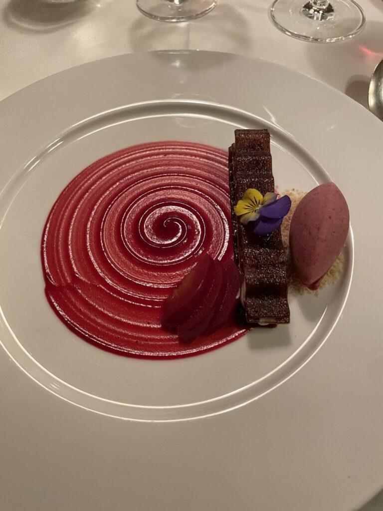 Ein Dessert auf einer Platte mit einer roten Spiralsauce, Rübenscheiben, einem rechteckigen Schokoladenkuchen, einer Kugel Beerensorbet und einer essbaren Blume auf einem weißen Teller.