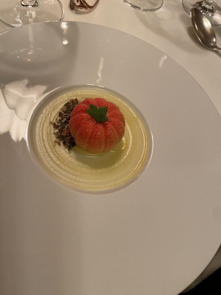 Eine Nachspeise in Form einer roten Tomate mit grünem Stiel, serviert auf einer gelben Sahnesauce und garniert mit Schokoladenstreuseln.