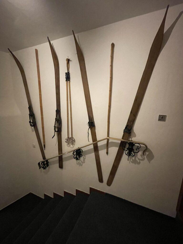 An einer weißen Wand über einer Treppe mit schmiedeeisernem Geländer sind alte Holzskier und Bambusskistöcke vertikal angebracht.