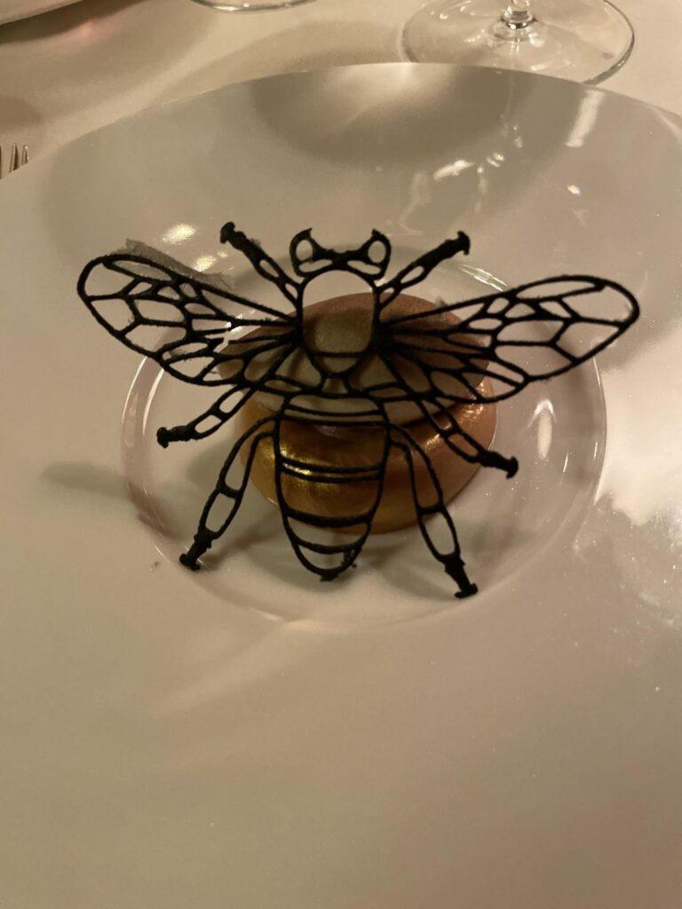 Ein Dessert auf einem weißen Teller mit einer verschlungenen, essbaren Dekoration in Form einer Biene, mit einer Gabel und einem Weinglas im Hintergrund.