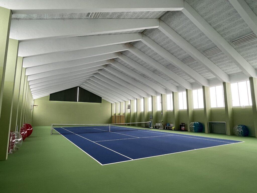 Tennishalle mit blauer Spielfläche und grüner Umgebung, unter einer hohen, weißen, schrägen Decke mit großen Fenstern, die Tageslicht hereinlassen.