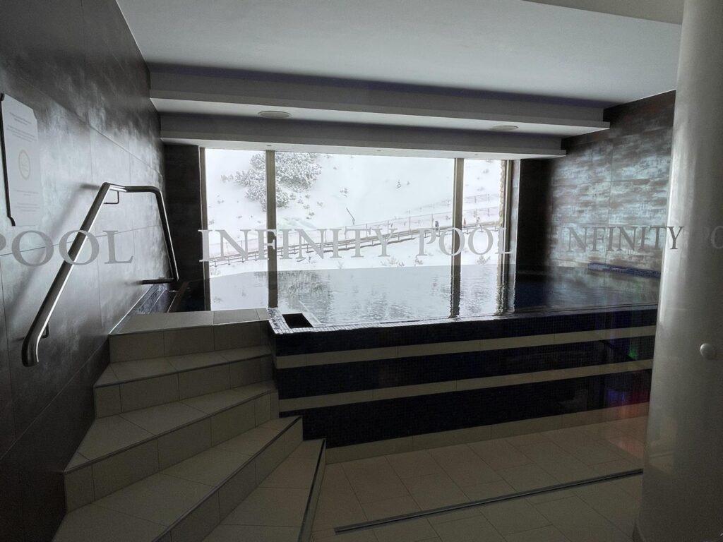 Indoor-Infinity-Pool mit Stufen zum Einstieg, dunklen Fliesenwänden und einem großen Fenster, das die verschneite Landschaft draußen zeigt.