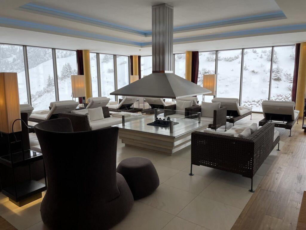 Modernes Wohnzimmer mit großen Fenstern, die eine verschneite Landschaft zeigen, mit zentralem Kamin, verschiedenen Sesseln und Sofas, die zum Entspannen einladen.