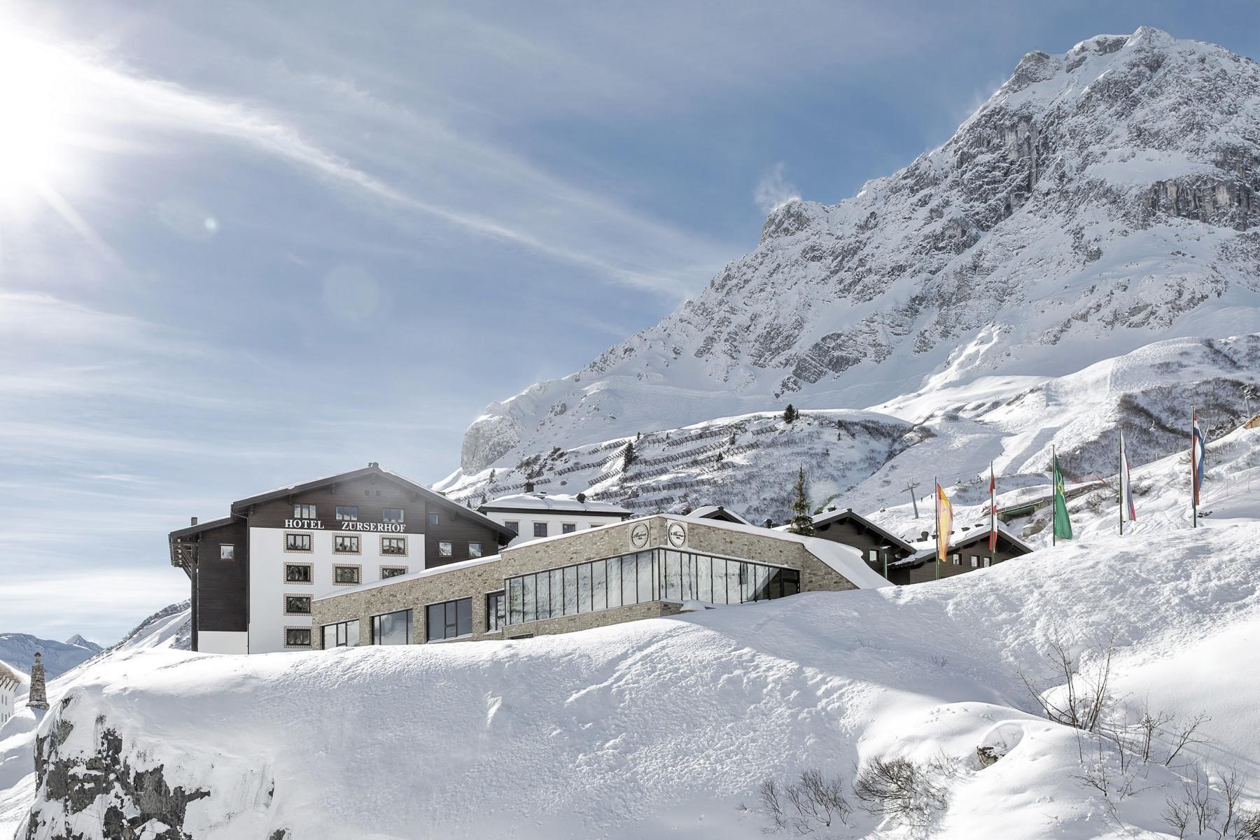 Ein Hotelgebäude ist von Schnee umgeben, vor dem Hintergrund schneebedeckter Berge und eines strahlend blauen Himmels; draußen sind mehrere Fahnen aufgestellt.