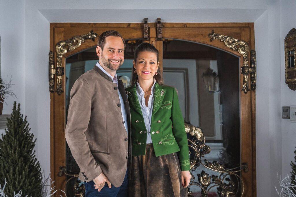 Ein Mann und eine Frau stehen lächelnd und in traditioneller österreichischer Kleidung vor einer verzierten Holztür.