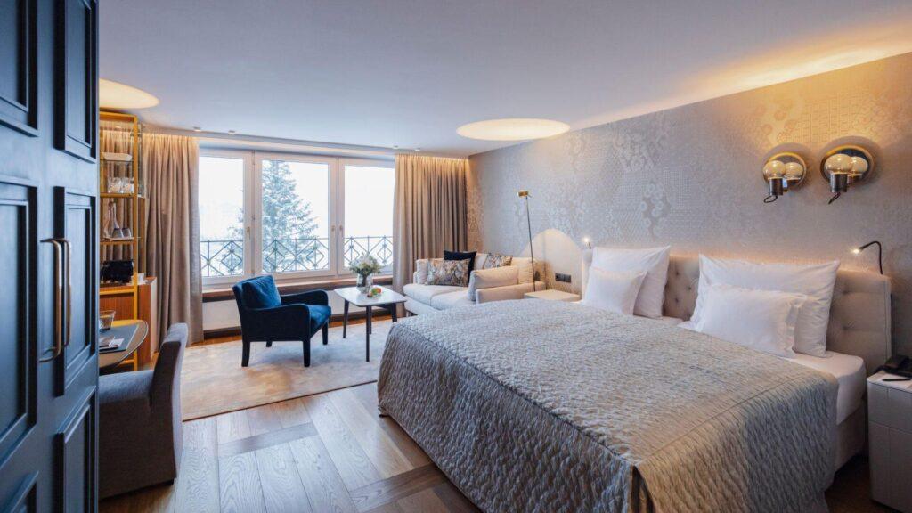 Modernes Hotelzimmer mit großem Bett, Sitzecke mit Stühlen und Sofa, Schreibtisch und großen Fenstern mit Blick auf eine verschneite Landschaft.