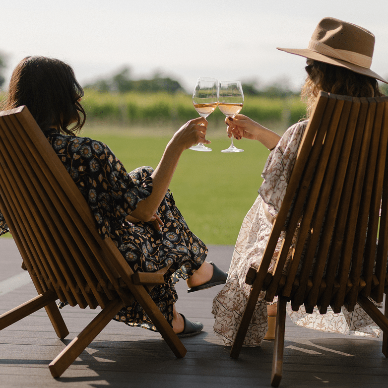 Zwei Frauen sitzen in Holzstühlen auf einer Terrasse im Freien und stoßen mit Weingläsern an, mit Blick auf grünes Gras und Bäume in der Ferne.
