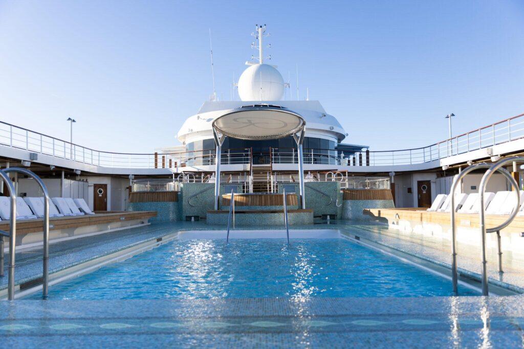 Ein klarer Swimmingpool mit Stufen führt zu einem modernen Kreuzfahrtschiffdeck mit Liegestühlen unter blauem Himmel.