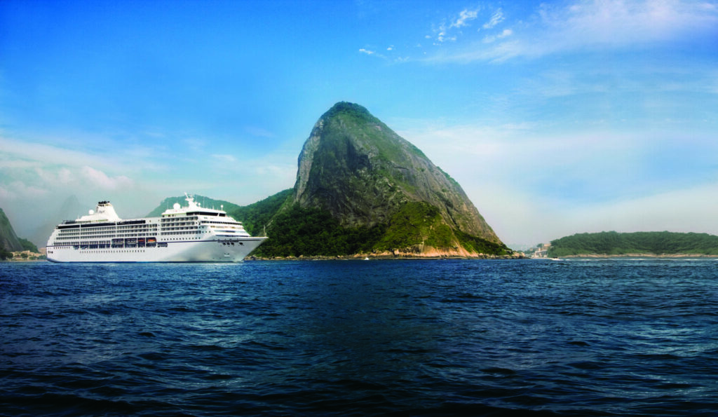 Ein weißes Kreuzfahrtschiff fährt in der Nähe eines großen, steilen Berges an der Küste von Rio de Janeiro in Brasilien unter blauem Himmel.