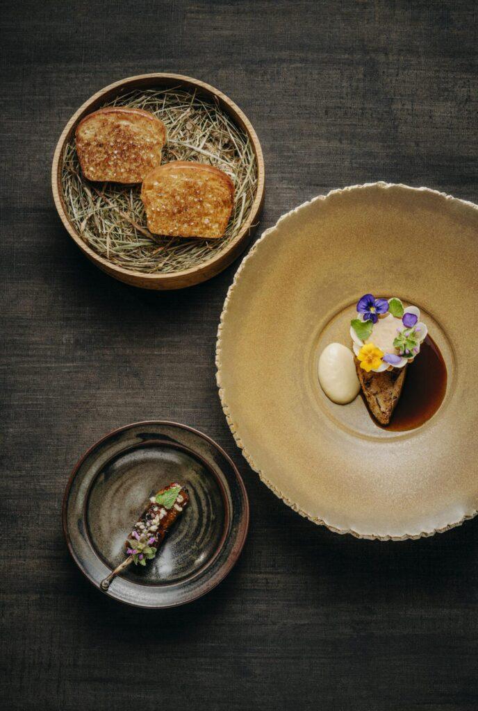 Drei Teller auf einer dunklen Fläche: zwei Stücke Brot auf Heu, ein dekoriertes Dessert mit Soße und Sahne und eine kleine Schokoladenleckerei, garniert mit Kräutern.