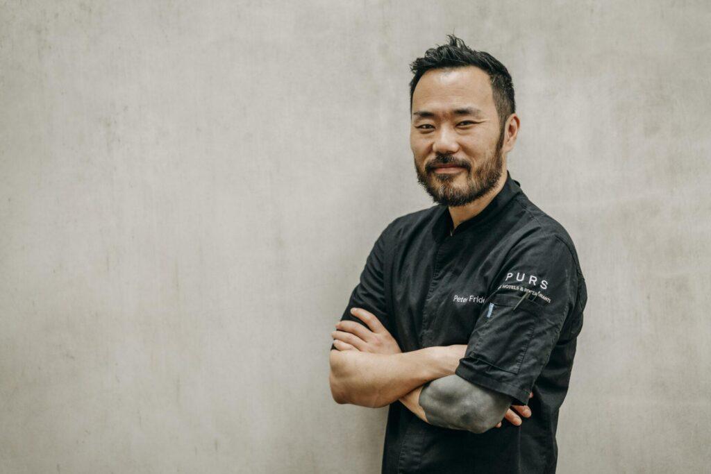 Ein Mann in einer schwarzen Kochjacke steht mit verschränkten Armen und einem leichten Lächeln vor einer schlichten hellen Wand.