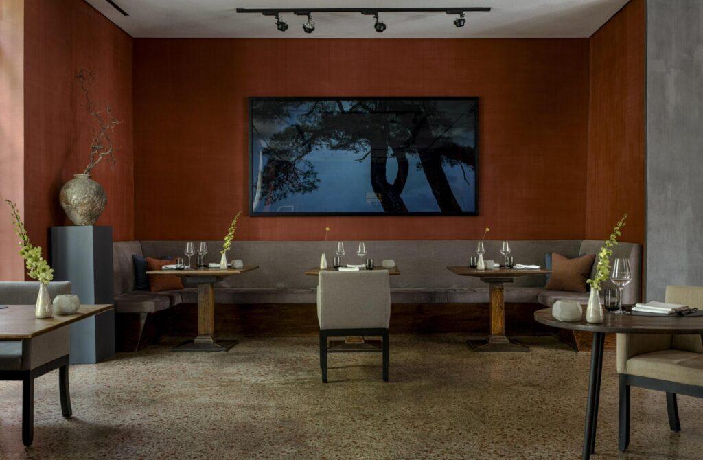 Ein modernes Restaurant mit braunen Wänden, minimalistisch gedeckten Tischen, gepolsterten Sitzbänken und einem großen gerahmten Kunstwerk über dem Sitzbereich.