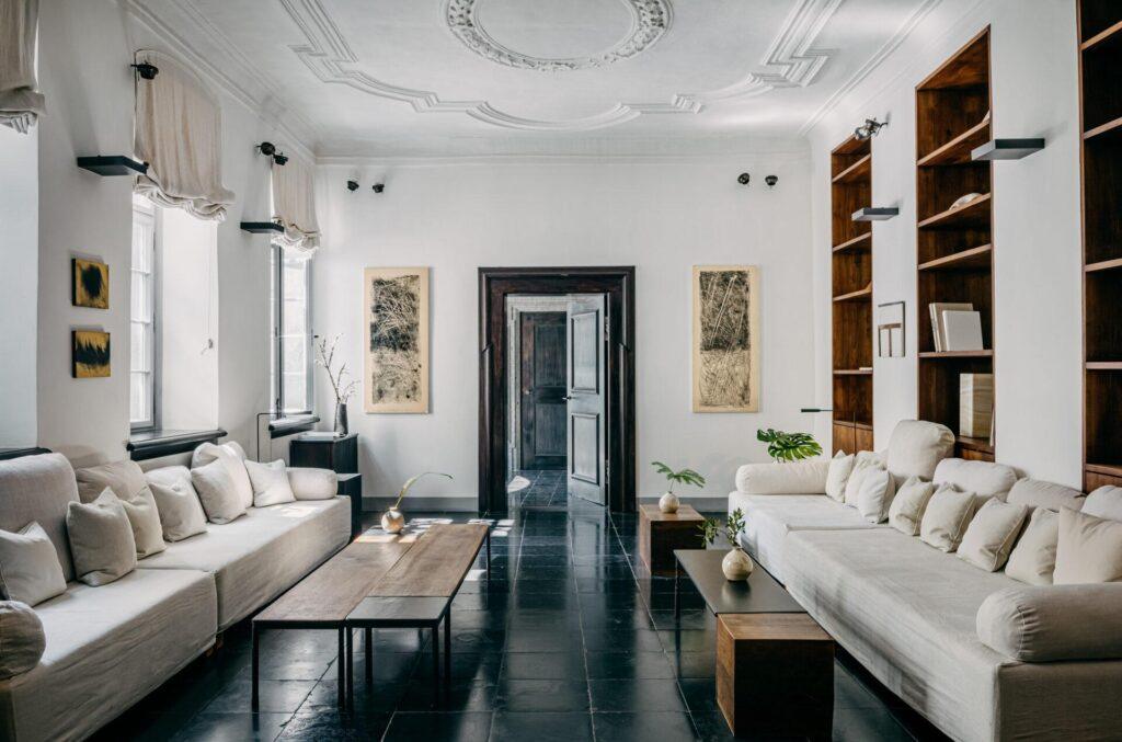 Ein geräumiges Wohnzimmer mit zwei weißen Sofas, einem Couchtisch aus Holz, Einbauregalen, schwarzen Fliesenböden und hohen Fenstern mit hellen Vorhängen.