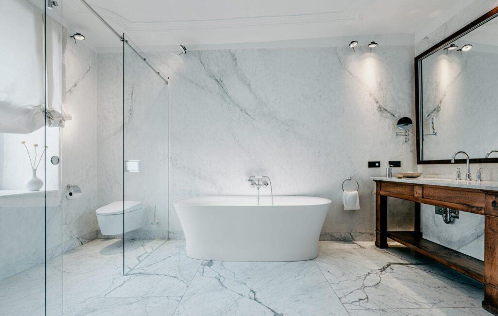 Modernes Badezimmer mit Marmorböden und -wänden, einer freistehenden Badewanne, einer Glasdusche, einer wandmontierten Toilette, einem großen hölzernen Waschtisch und einem gerahmten Spiegel.