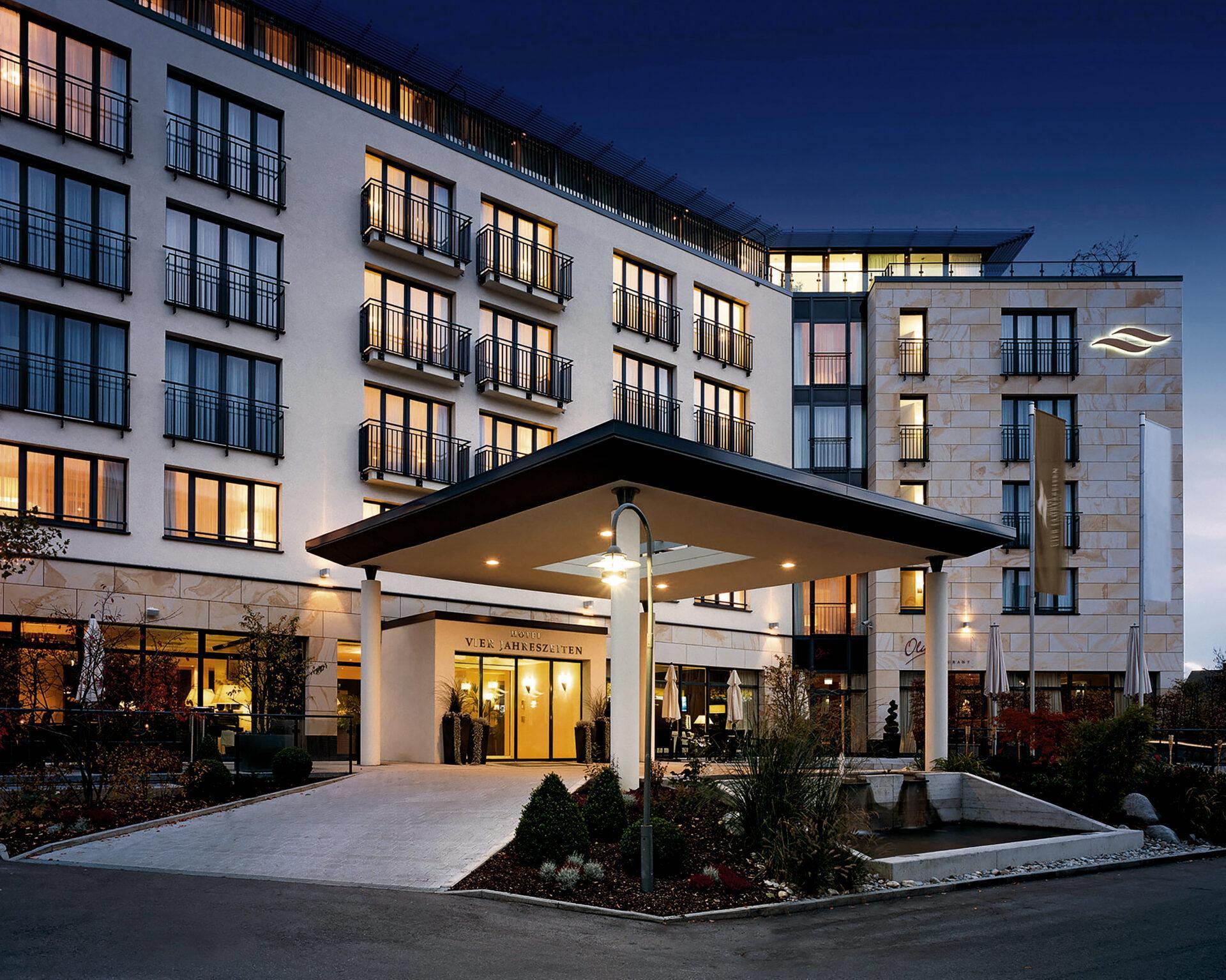 Modernes Hotelgebäude mit mehreren Etagen und Balkonen, das in der Abenddämmerung beleuchtet wird, mit überdachtem Eingang und begrünter Umgebung.