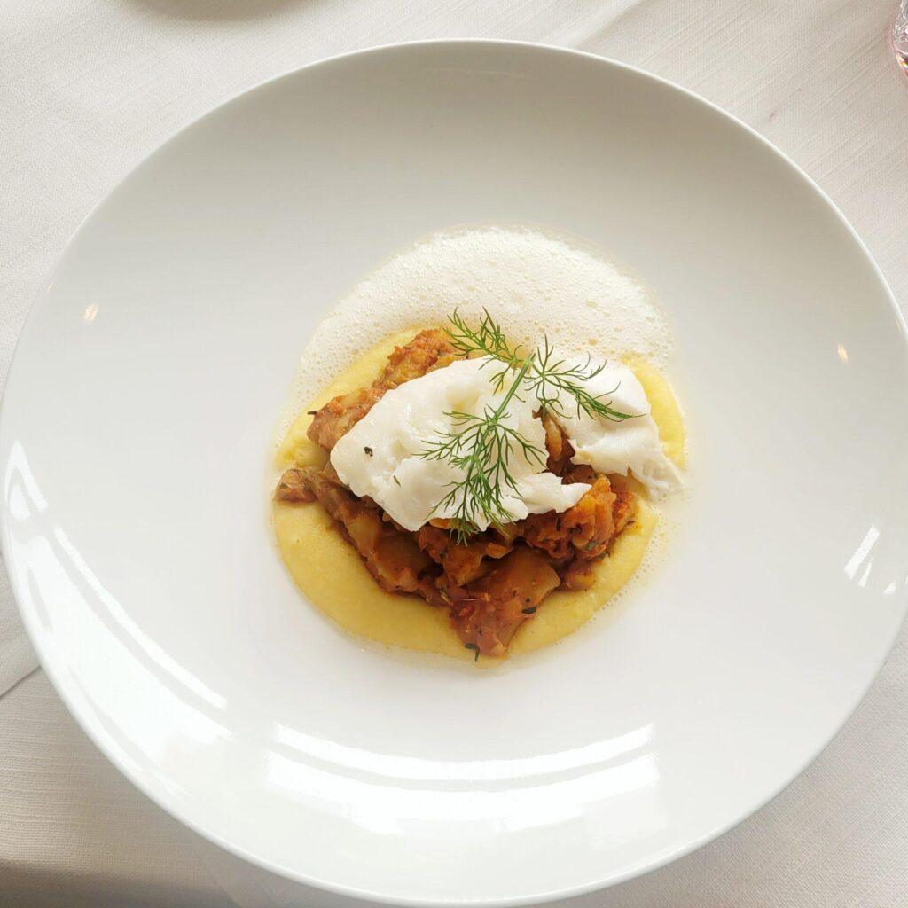 Ein Tellergericht mit einer Portion pochiertem Weißfisch auf einem Bett aus cremigem gelbem Püree, garniert mit Dill und leichtem Schaum, serviert auf einem weißen runden Teller.