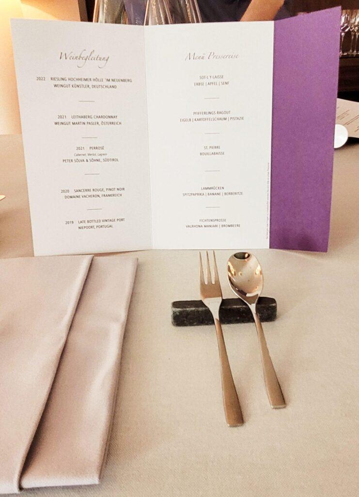 Eine gefaltete Speisekarte mit Weinkarte und Menüvorschau steht auf einem Tisch, der mit Serviette, Gabel, Löffel und Messerablage gedeckt ist.