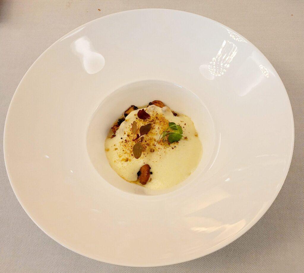 Eine weiße Schale mit einem Gourmetteller mit cremigem Schaum, garniert mit Kräutern und kleinem Gemüse, auf einem hellen Tischtuch.