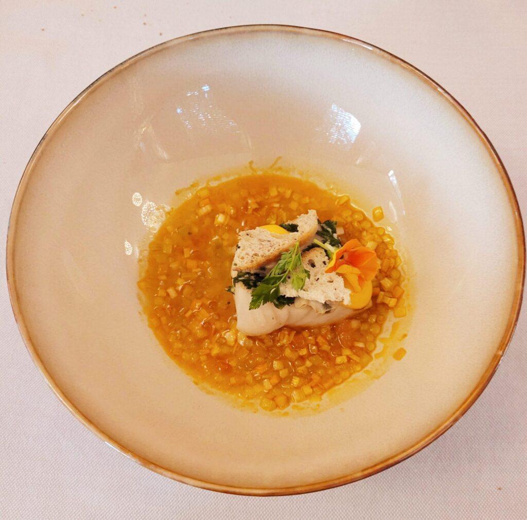 Ein Fisch mit gewürfeltem Gemüse in einer gelben Brühe, dazu Kräuter und ein knuspriges Brot auf einem runden Teller.