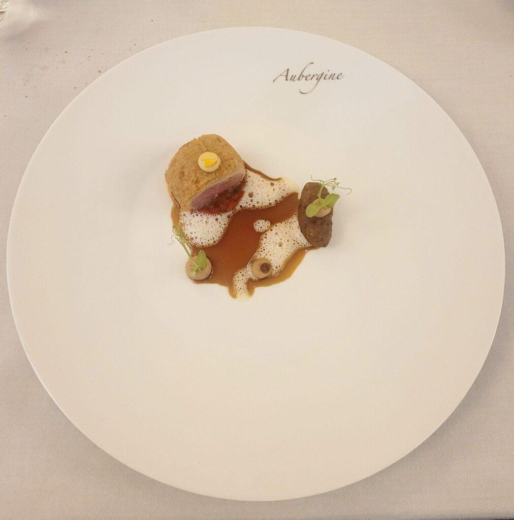 Ein minimalistisches Gourmet-Gericht mit einer kleinen Portion Fleisch, eingewickelt in Teig, Schaum, Sauce und Beilage, präsentiert auf einem großen weißen Teller mit der Aufschrift "Aubergine".