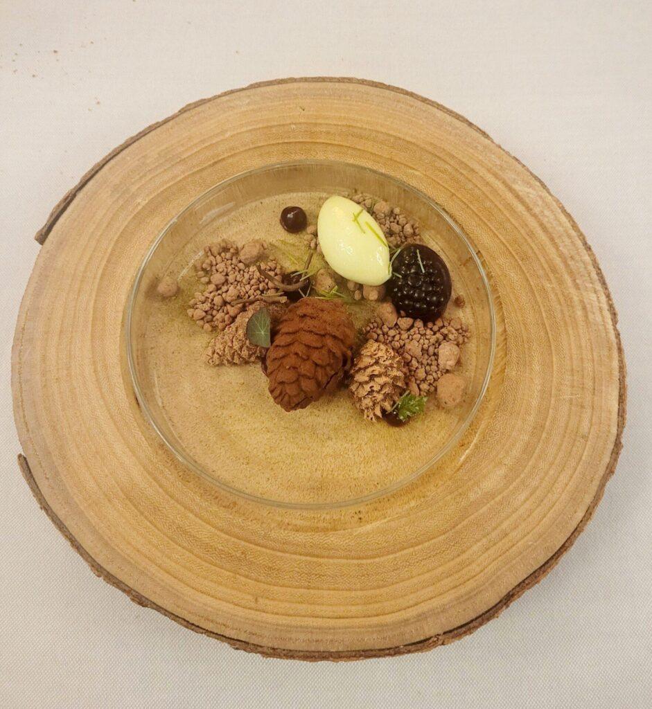 Ein Dessert in Form einer Waldszene mit Schokoladenstücken in Form von Tannenzapfen und Eis, serviert auf einer Glasplatte auf einer Holzplatte.