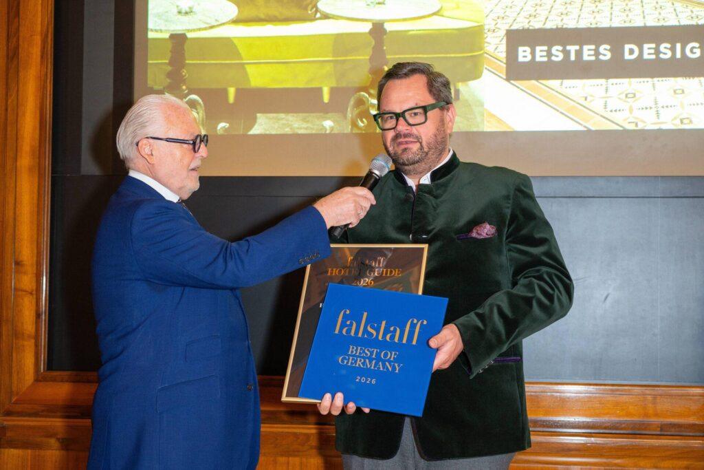 Zwei Männer stehen in einem Innenraum; einer hält dem anderen ein Mikrofon hin, der eine Plakette und ein blaues Preisschild mit der Aufschrift "falstaff Best of Germany 2024" hält. Im Hintergrund ist eine Präsentationsleinwand zu sehen.