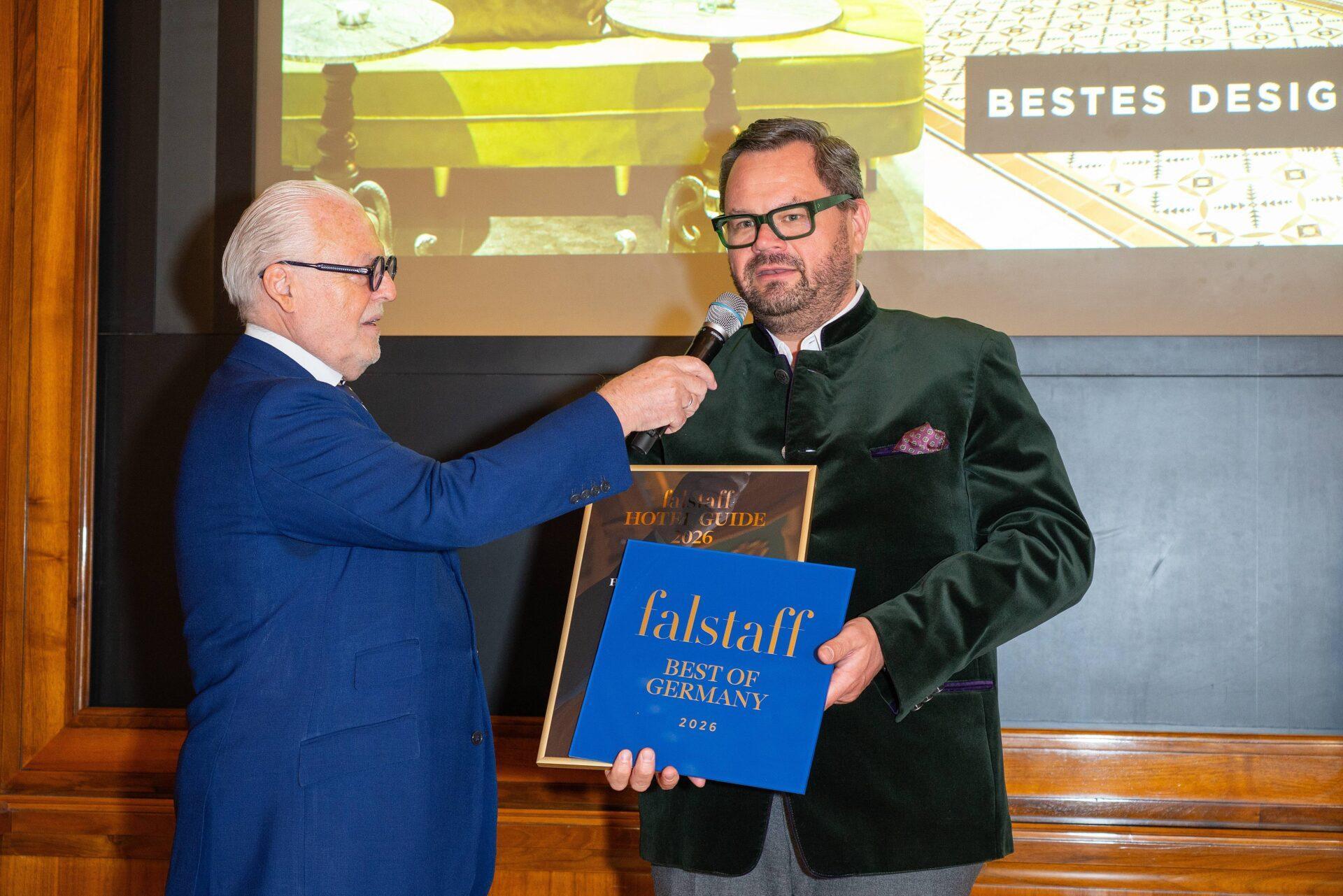 Zwei Männer stehen in einem Innenraum; einer hält dem anderen ein Mikrofon hin, der eine Plakette und ein blaues Preisschild mit der Aufschrift "falstaff Best of Germany 2024" hält. Im Hintergrund ist eine Präsentationsleinwand zu sehen.