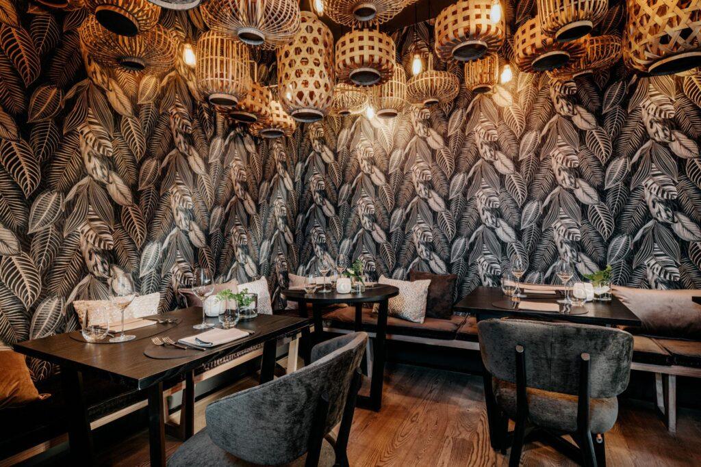 Ein kleiner Restaurantbereich mit dunklen Holzmöbeln, botanisch gemusterten Tapeten und hängenden Korbleuchten.