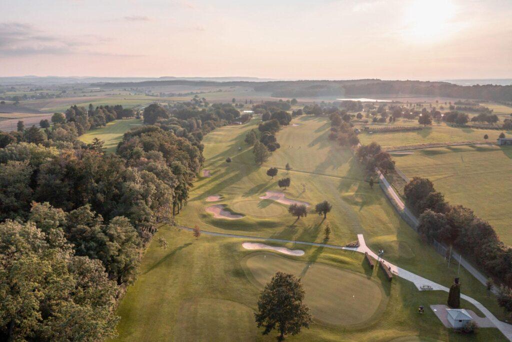 Luftaufnahme eines Golfplatzes mit mehreren Grüns, Sandbunkern, Bäumen und verschlungenen Wegen unter einem teilweise bewölkten Himmel bei Sonnenuntergang.